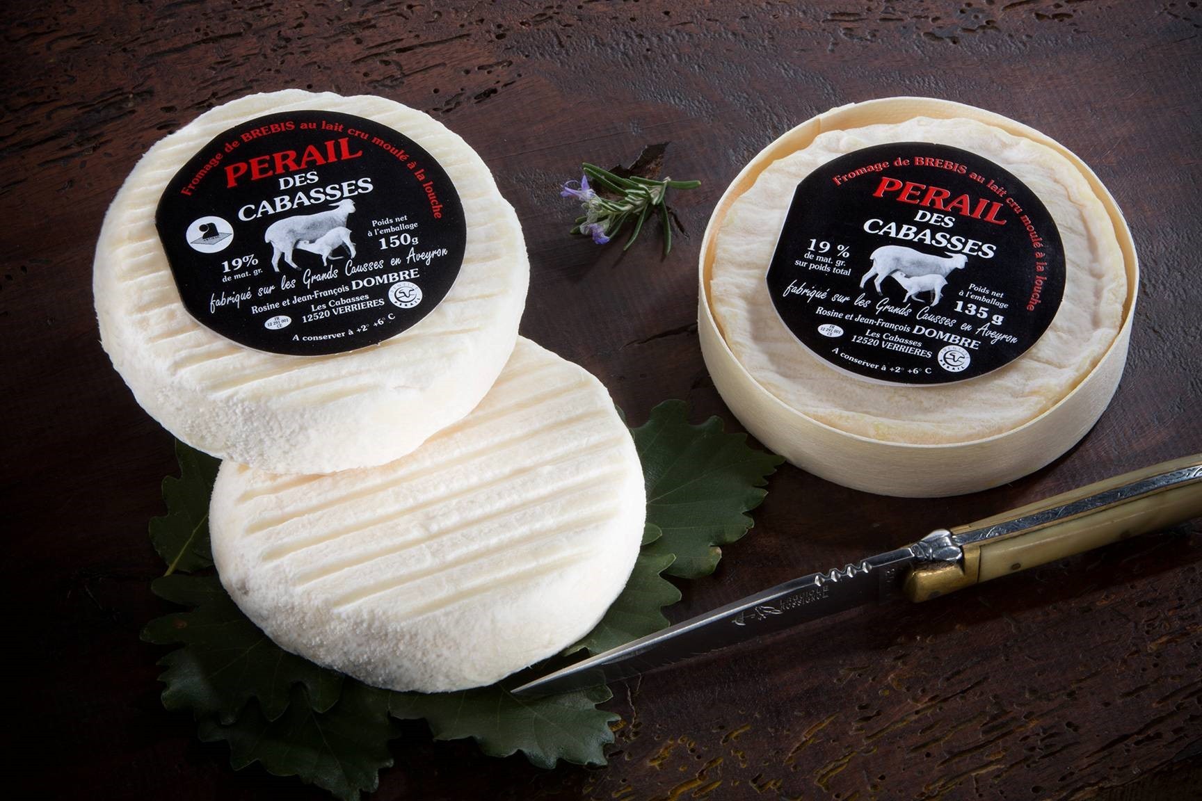 Deux meules rondes de fromage Pérail des Cabasses aux étiquettes noires reposent sur une surface en bois à côté d'un couteau, de feuilles vertes et d'une petite fleur violette.