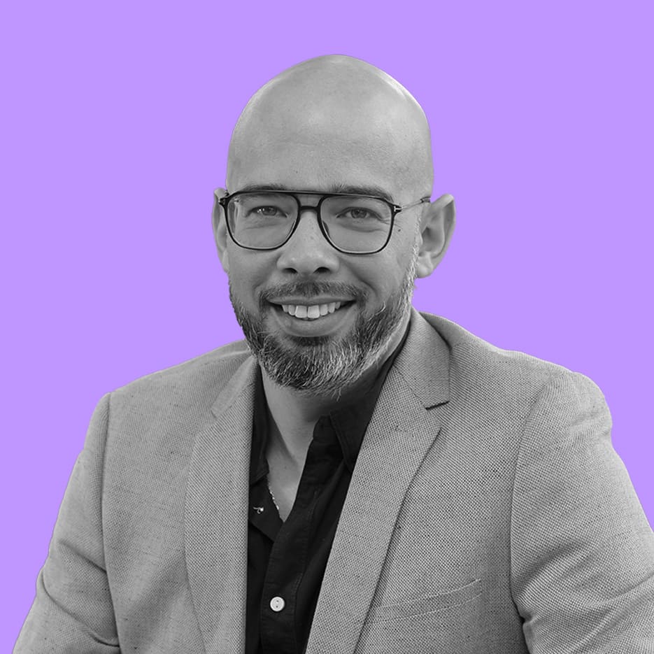 Un homme chauve avec des lunettes et une barbe courte, habillé en courtisan en énergie avec un blazer clair et une chemise foncée, sourit à la caméra sur un fond violet uni.