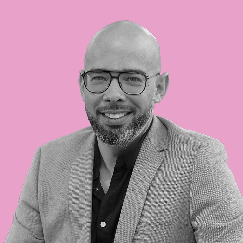 Un gestionnaire de réseau portant des lunettes, une tête chauve et une barbe porte un blazer clair et une chemise foncée, posant sur un fond rose uni.