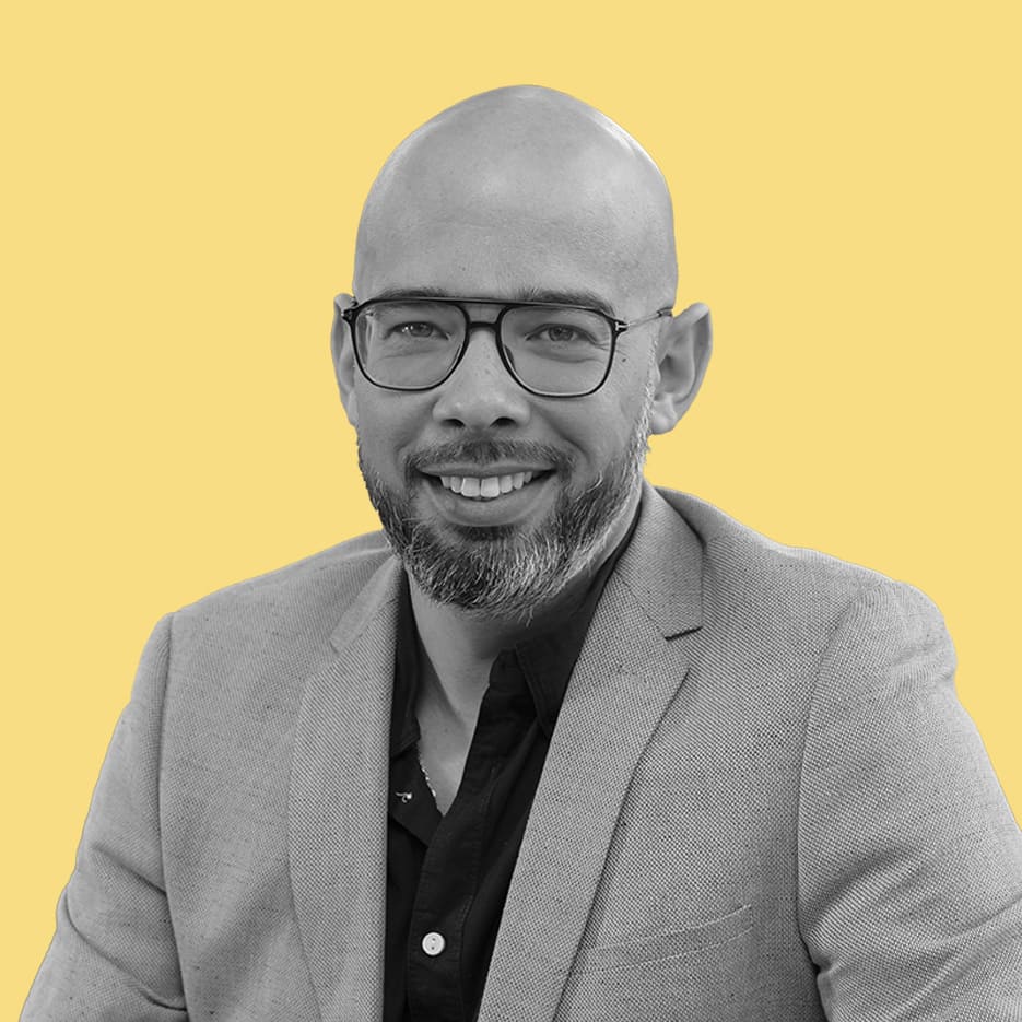 Un homme chauve avec des lunettes et une barbe, vêtu d'un blazer clair sur une chemise foncée, sourit à la caméra sur un fond jaune uni, représentant un cabinet de courtage en expansion.