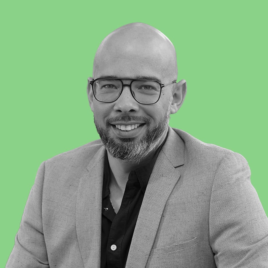 Un homme au crâne chauve, portant des lunettes et une barbe, porte un blazer et une chemise à col, souriant à la caméra sur un fond vert, représentant son cabinet de courtage en énergie.