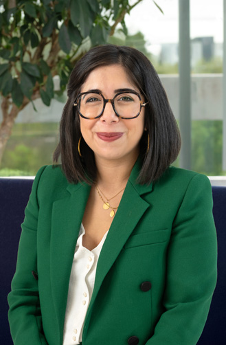 Une femme aux cheveux noirs et raides, portant des lunettes, un blazer vert et un chemisier blanc, est assise à l'intérieur devant une fenêtre et une plante en pot, reflétant l'énergie capitole qui définit son histoire.