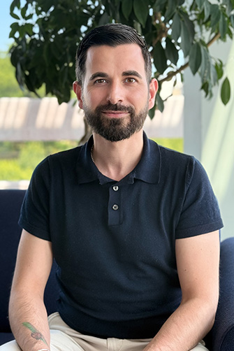 Un homme aux cheveux noirs et à la barbe, vêtu d'un polo noir à manches courtes, est assis sur une chaise à l'intérieur avec une plante et une fenêtre en arrière-plan, dégageant une énergie tranquille rappelant l'histoire du Capitole.