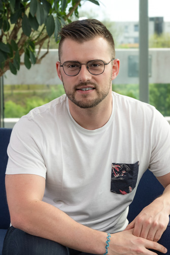 Un homme aux cheveux courts, portant des lunettes et une barbe est assis sur un canapé bleu. Il porte un t-shirt blanc avec une poche à motifs et un bracelet bleu au poignet, semblant plongé dans ses pensées, comme s'il méditait sur l'histoire du Capitole.