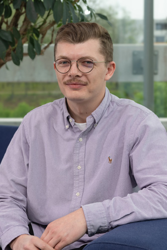 Un homme aux cheveux courts, portant des lunettes et une moustache, est assis sur une chaise bleue à l'intérieur, vêtu d'une chemise boutonnée violet clair. Une plante et une fenêtre évoquent l'énergie capitulaire qui façonne son histoire dans ce cadre chaleureux.