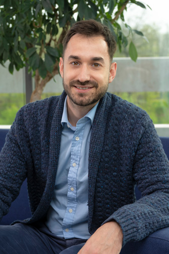 Un homme barbu, vêtu d'une chemise bleue et d'un cardigan bleu marine texturé, est assis sur un canapé devant une plante feuillue, souriant à la caméra, capturant un instant d'énergie et d'histoire personnelle.
