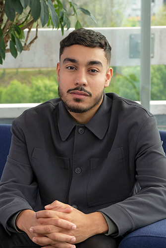 Un homme aux cheveux noirs courts et à la barbe taillée est assis sur un canapé bleu, vêtu d'une veste sombre, les mains jointes et l'air neutre. Une plante dynamise l'arrière-plan, évoquant son histoire personnelle.