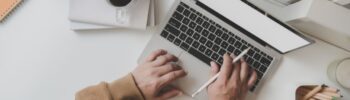 Une personne travaillant sur un ordinateur portable avec un stylet, concentrée sur l'approvisionnement en énergie de l'entreprise, est assise à côté d'une tasse de café, d'un cahier et d'un contenant de crayons de couleur sur un bureau blanc propre.
