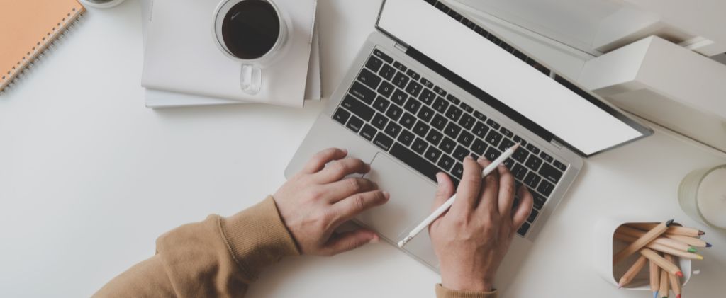 Une personne travaillant sur un ordinateur portable avec un stylet, concentrée sur l'approvisionnement en énergie de l'entreprise, est assise à côté d'une tasse de café, d'un cahier et d'un contenant de crayons de couleur sur un bureau blanc propre.