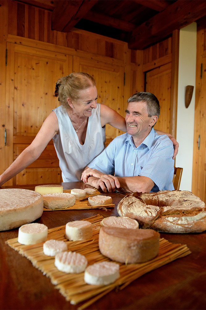 Une femme et un homme se sourient dans une cuisine en bois, avec divers fromages ronds et des pains disposés sur la table devant eux.