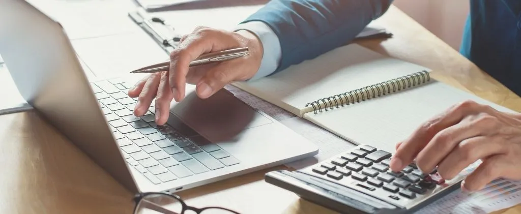 Personne travaillant à un bureau avec un ordinateur portable, une calculatrice et un bloc-notes, examinant des documents et tenant un stylo, calculant peut-être les coûts de la taxe d'électricité.