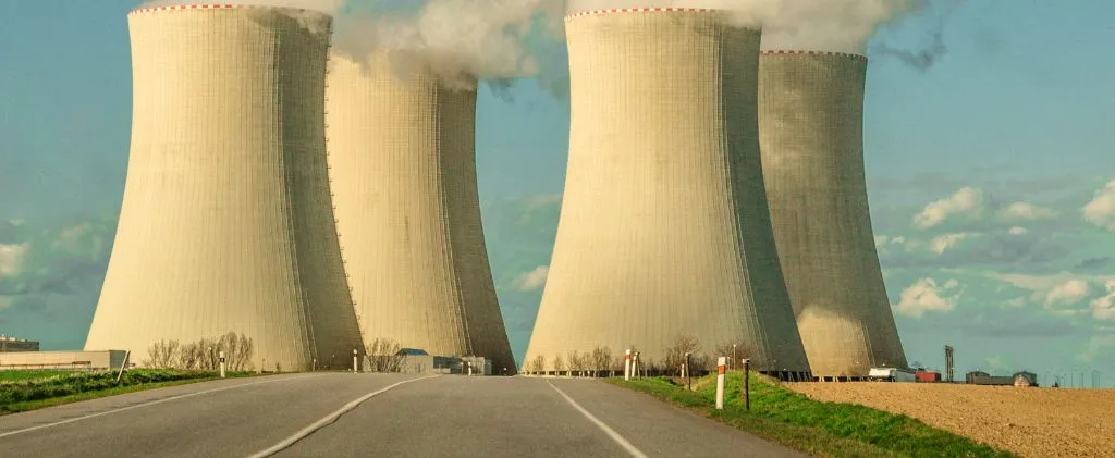 Quatre grandes tours de refroidissement d'une centrale nucléaire émettent de la vapeur, vues depuis une route menant à l'installation sous un ciel partiellement nuageux, soulignant l'ampleur de la production d'énergie.