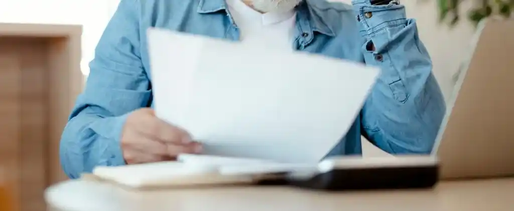 Une personne portant une chemise en jean examine des documents – probablement sur la taxe électrique – à un bureau avec un ordinateur portable, un carnet et un objet noir au premier plan.