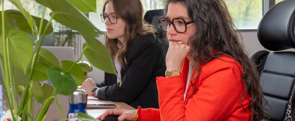 Deux femmes portant des lunettes travaillent à des bureaux équipés d'ordinateurs ; l'une d'elles, vêtue d'une veste rouge, se concentre sur son écran, analysant peut-être des données de taxe électricité, tandis que des plantes vertes ajoutent de la fraîcheur à l'espace de travail au premier plan.