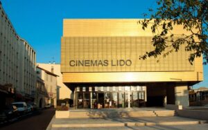 Un bâtiment de cinéma moderne avec une grande façade dorée affichant le panneau « CINEMAS LIDO » au-dessus des portes d'entrée en verre, sous un ciel bleu clair.
