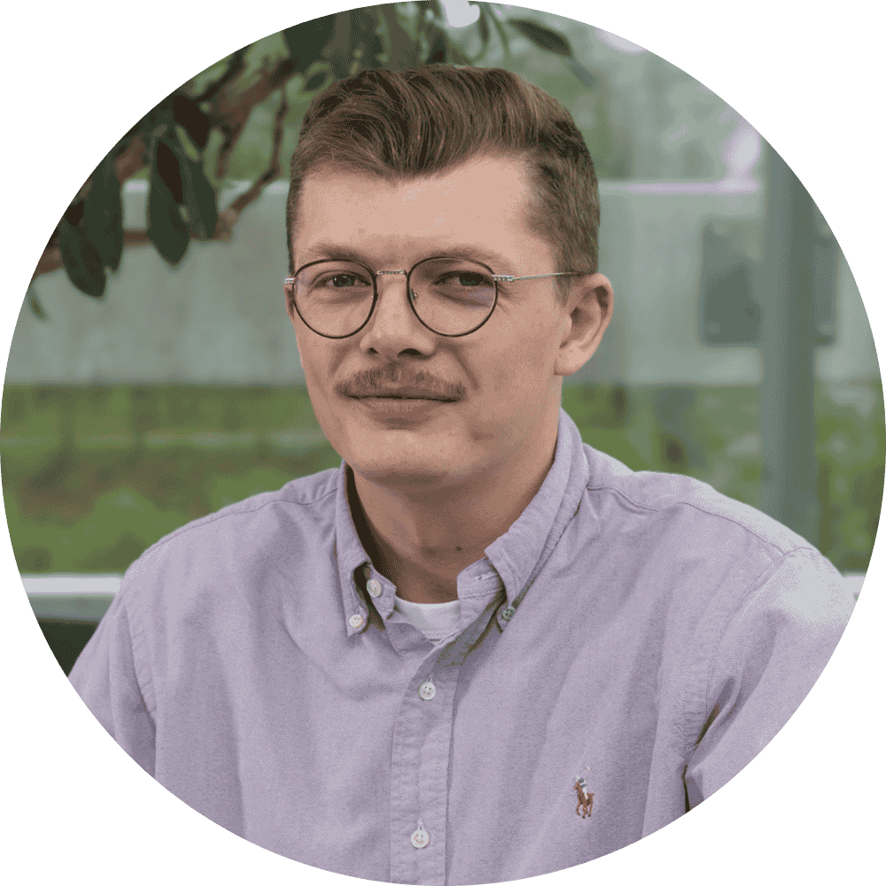 Un homme aux cheveux bruns courts, aux lunettes et à la moustache porte une chemise boutonnée violet clair et est assis à l'intérieur avec de la verdure visible en arrière-plan.