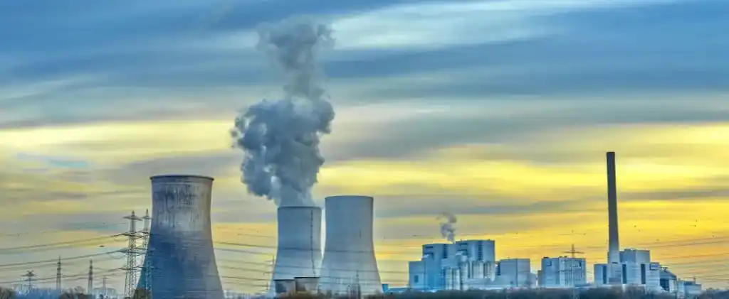 Une centrale électrique avec des tours de refroidissement émettant de la vapeur et de la fumée sous un ciel nuageux au coucher du soleil, entourée d'infrastructures électriques, se dresse alors que la nouvelle réglementation TURPE 7 impacte le paysage énergétique.