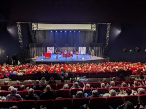 Un public de théâtre assiste à une représentation sur scène avec des décorations de Noël et des artistes sous un éclairage vif.