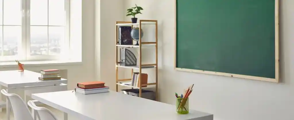 Une salle de classe avec des bureaux blancs, des livres empilés, des chaises, un tableau vert et une étagère décorative bénéficie de la lumière du soleil à travers de grandes fenêtres - un exemple de décret tertiaire bâtiments scolaires conformité pour l'efficacité énergétique.