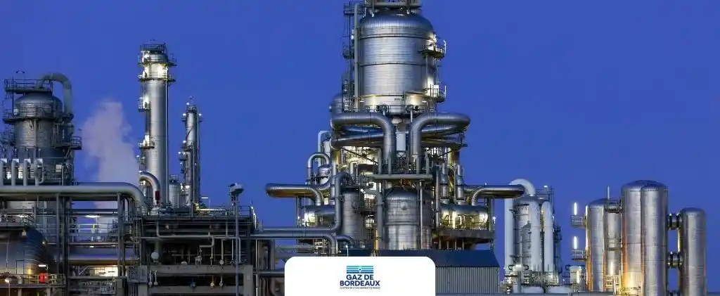 Usine de gaz industriel avec de grands réservoirs métalliques et des tuyaux sous un ciel clair en soirée ; un panneau "GAZ DE BORDEAUX PRO" est placé bien en vue à l'avant de l'installation.
