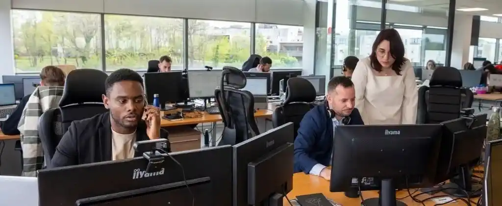 Environnement de bureau avec plusieurs personnes travaillant à des bureaux équipés d'ordinateurs ; un courtier en énergie est au téléphone, un autre tape, et une femme se tient à côté de lui en regardant son moniteur, lançant des défis aux idées reçues sur les solutions en matière d'énergie.