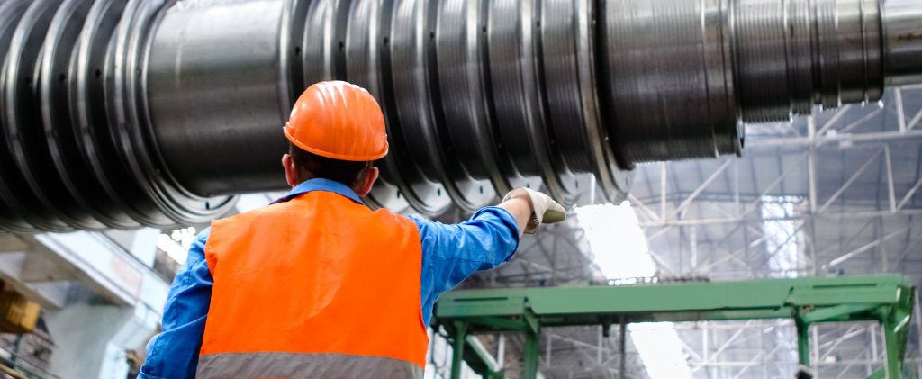 Un travailleur vêtu d'un gilet de sécurité et d'un casque orange inspecte de grosses machines industrielles à l'intérieur d'une usine dotée de structures métalliques et d'un éclairage aérien, semblable à la précision opérationnelle que l'on trouve au Capitole Énergie et Les Cinémas de Saint-Raphaël.