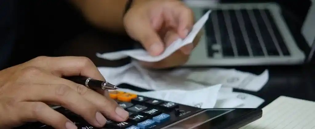 Une personne utilise une calculatrice et tient des reçus tout en travaillant sur un ordinateur portable, avec un bloc-notes à proximité, examinant des factures liées à tva énergie 2025.