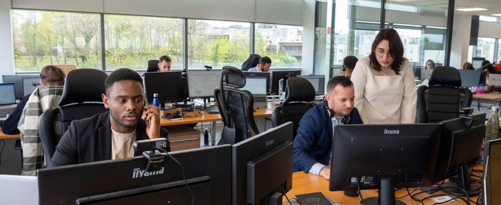 Environnement de bureau avec plusieurs personnes travaillant à des bureaux équipés d'ordinateurs ; un courtier en énergie est au téléphone, un autre tape, et une femme se tient à côté de lui en regardant son moniteur, lançant des défis aux idées reçues sur les solutions en matière d'énergie.