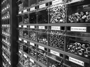 Dans une quincaillerie, des bacs en plastique transparent remplis d'écrous et de boulons métalliques de différentes tailles sont rangés sur des étagères étiquetées.