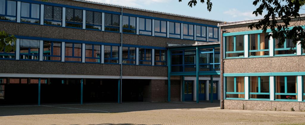 Un bâtiment scolaire moderne avec des cadres de fenêtres bleus, de grandes fenêtres pour l'économie d'énergie, et une cour pavée à l'avant, photographié par une journée ensoleillée.