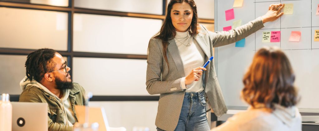 Une femme se tient près d'un tableau blanc couvert de notes adhésives. Elle tient un stylo et présente à deux collègues assis dans un bureau un exposé sur l'optimisation des puissances de raccordement.