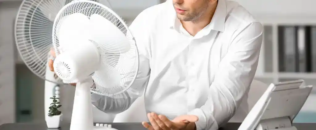 Un homme en chemise blanche est assis à un bureau, inspectant un ventilateur électrique blanc, explorant les options pour un achat dynamique énergie, avec un ordinateur portable et une petite plante à proximité.