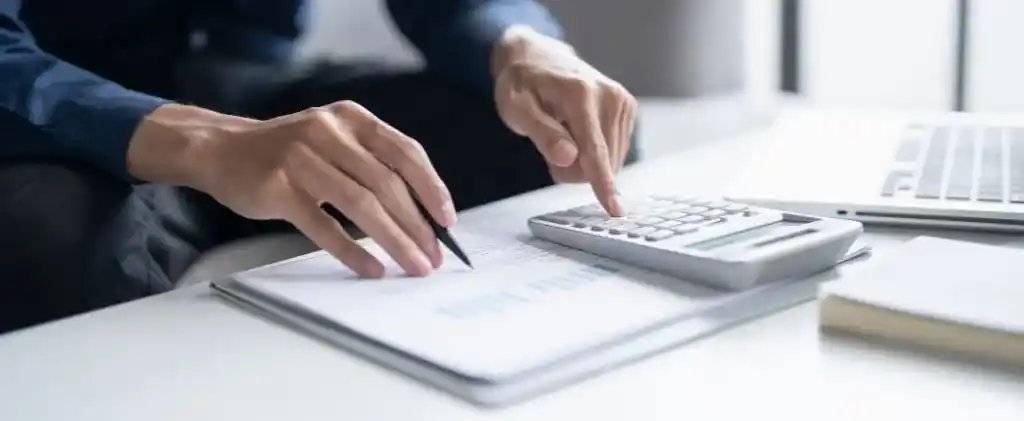 Une personne utilise une calculatrice tout en examinant des documents financiers à un bureau avec un ordinateur portable, cherchant des moyens de payer moins cher l'électricité.