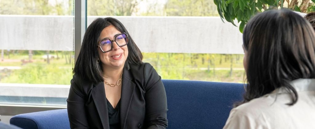 Une femme portant des lunettes et un blazer noir est assise sur un canapé bleu, souriant et engageant une conversation sociale avec une autre personne à l'intérieur, près d'une fenêtre.