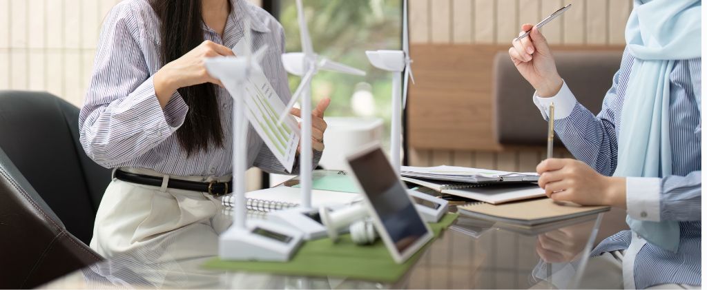 Deux personnes sont assises à un bureau et discutent de documents et de modèles d'éoliennes, avec des ordinateurs portables, des blocs-notes et des stylos visibles sur la table, alors qu'elles explorent l'impact sociétal positif des solutions d'énergie renouvelable.