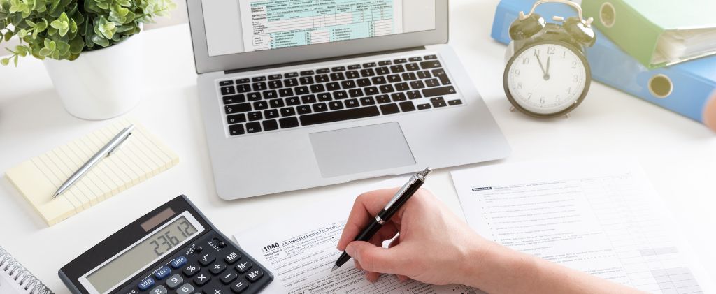 Une personne remplit des formulaires fiscaux à un bureau avec un ordinateur portable, une calculatrice, un stylo, une horloge, des documents et une petite plante en pot, tout en travaillant sur un projet RSE.