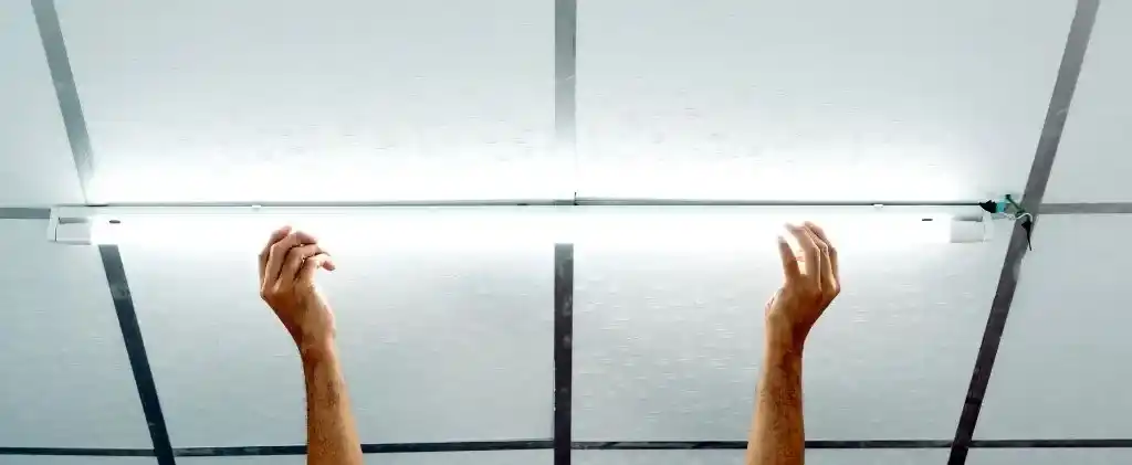Deux mains se tendent pour ajuster ou installer un luminaire fluorescent sur un plafond à dalles carrées, mettant en évidence des alternatives pratiques de climatisation bureau pour améliorer le confort et l'efficacité des bureaux.