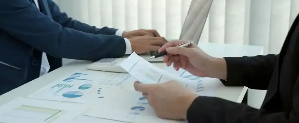 Deux personnes en tenue de travail examinent des graphiques et des tableaux à un bureau, analysant le coût du nucléaire, l'une utilisant un clavier et l'autre tenant des papiers et un stylo.