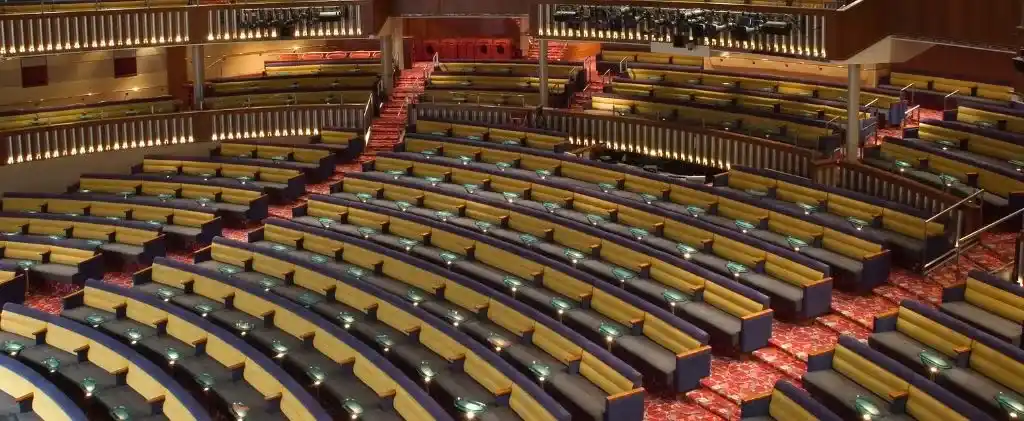 Un grand auditorium vide avec des rangées incurvées de sièges jaunes et bleus, une moquette à motifs rouges et des balcons de chaque côté, avec une signalisation claire pour le respect de l'EPI 3 retard.