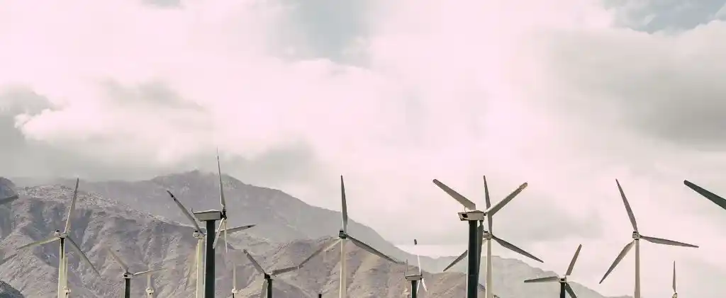 Des éoliennes se dressent devant des montagnes sous un ciel nuageux, soulignant les efforts en matière d'énergie renouvelable, cruciaux pour la conformité au champ d'application 2 du protocole sur les émissions de gaz à effet de serre.