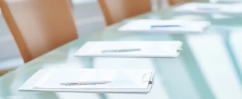 Gros plan d'une table de conférence avec des documents bien rangés, des rapports mettant en évidence le prix garantie d'origine, et des stylos placés devant des chaises bronzées dans une salle de réunion lumineuse.