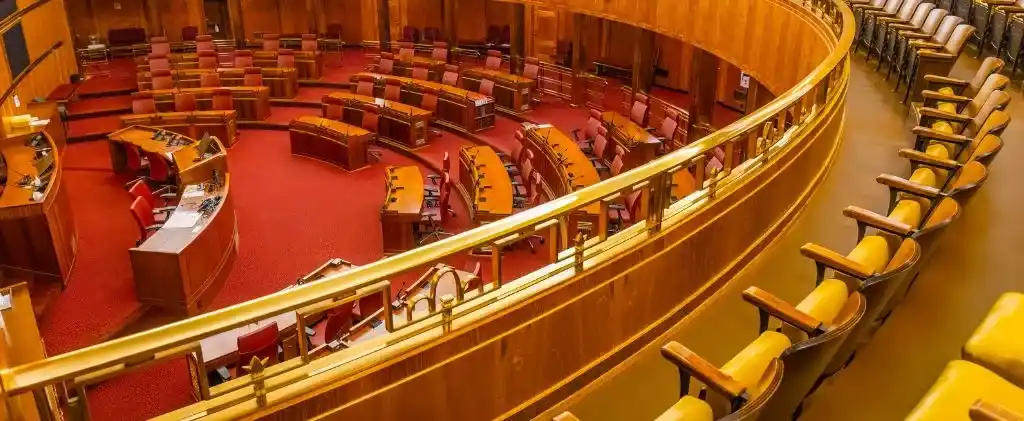 Une salle du gouvernement vide avec des rangées incurvées de bureaux en bois et une moquette rouge, vue d'en haut derrière des sièges de galerie à coussin jaune - une scène tranquille où des débats sur des sujets tels que le coût du nucléaire pourraient bientôt se dérouler.