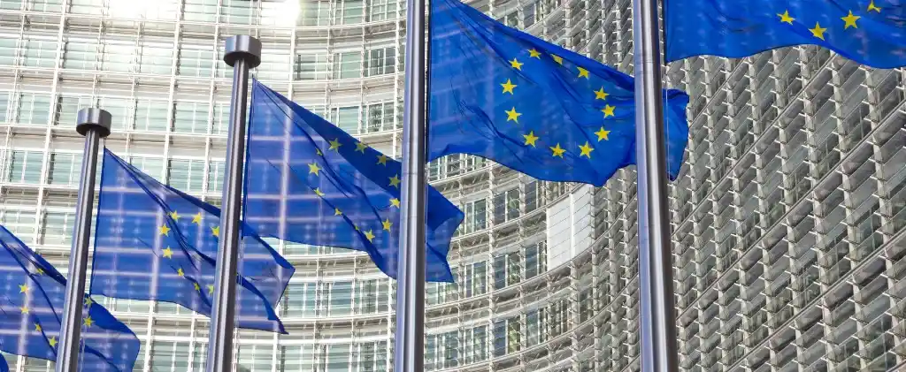 Plusieurs drapeaux de l'Union européenne flottent sur des mâts devant un immeuble de bureaux moderne en verre, illustrant les discussions sur la distribution du tarif gaz 2026 dans les États membres.