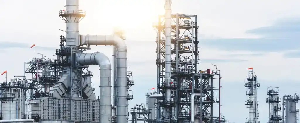 Un complexe de raffineries industrielles avec de nombreuses structures métalliques hautes, des tuyaux et des cheminées d'échappement se détachent sur un ciel nuageux.