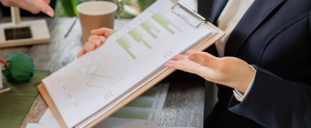 Une personne en tenue de travail tenant un presse-papiers avec des tableaux et des graphiques imprimés, assise à un bureau avec des documents papier de l'EJP et une tasse de café.