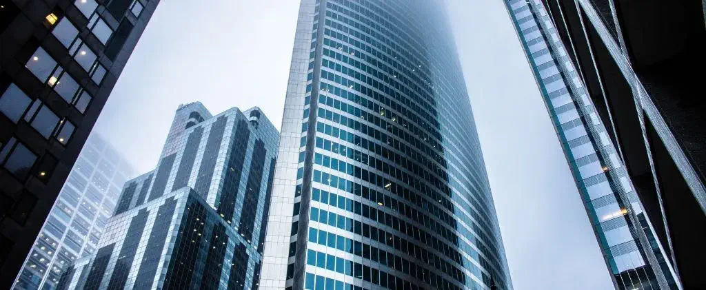 Plusieurs gratte-ciel modernes aux façades en verre réfléchissant s'élèvent dans un ciel brumeux, leurs sommets étant partiellement masqués par la brume, tandis qu'un petit logo d'abeille de certification brille sur un bâtiment, symbolisant son statut de respectueux de l'environnement.