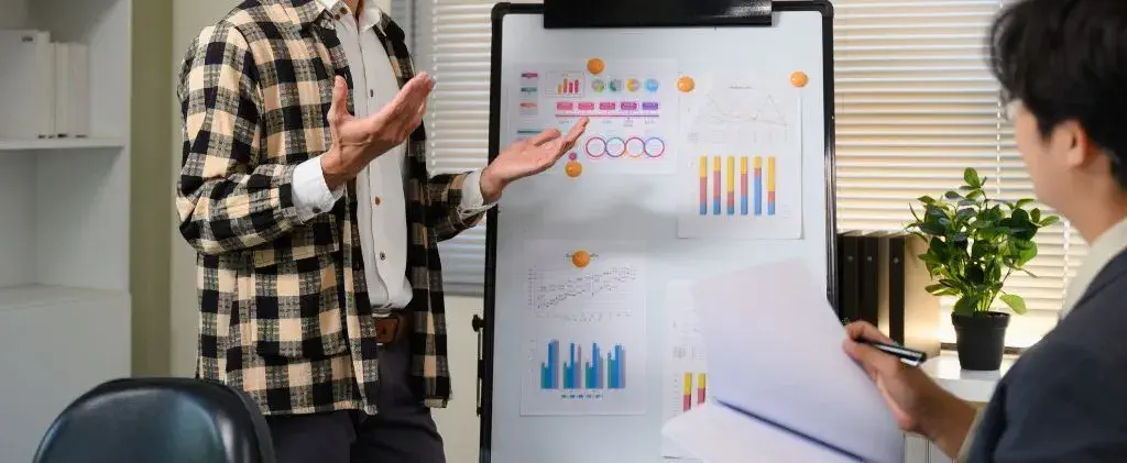 Deux personnes discutent d'un contrat d'électricité indexé dans un bureau ; l'une se tient près d'un tableau blanc avec des tableaux et des graphiques, tandis que l'autre est assise et prend des notes.