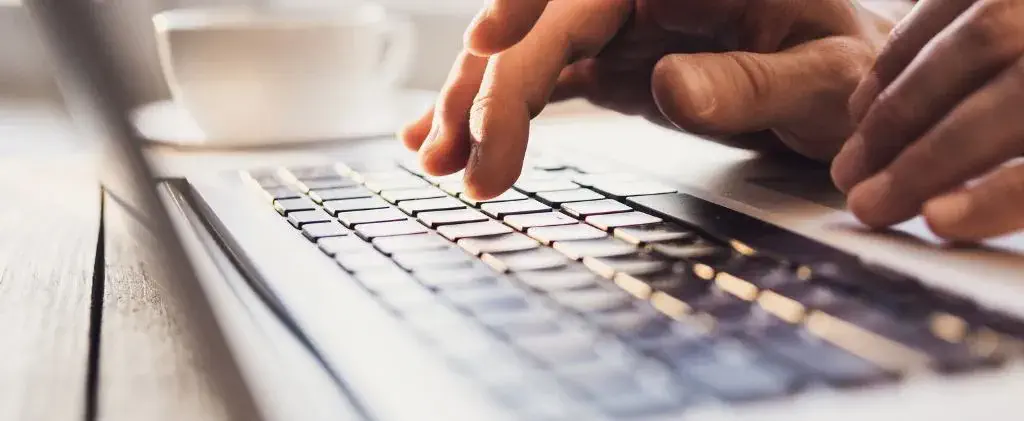Gros plan de mains tapant sur le clavier d'un ordinateur portable, recherchant des options pour un contrat d'électricité indexé, avec une tasse à café blanche en arrière-plan flou.