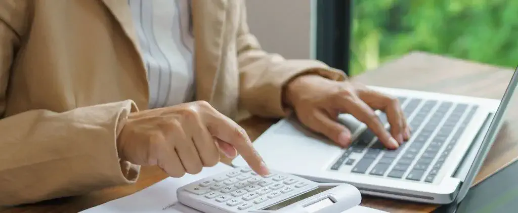 Une personne en veste beige révise les options prix fixe ou indexé, utilise une calculatrice et un ordinateur portable à un bureau, avec des papiers et une fenêtre montrant de la verdure en arrière-plan.