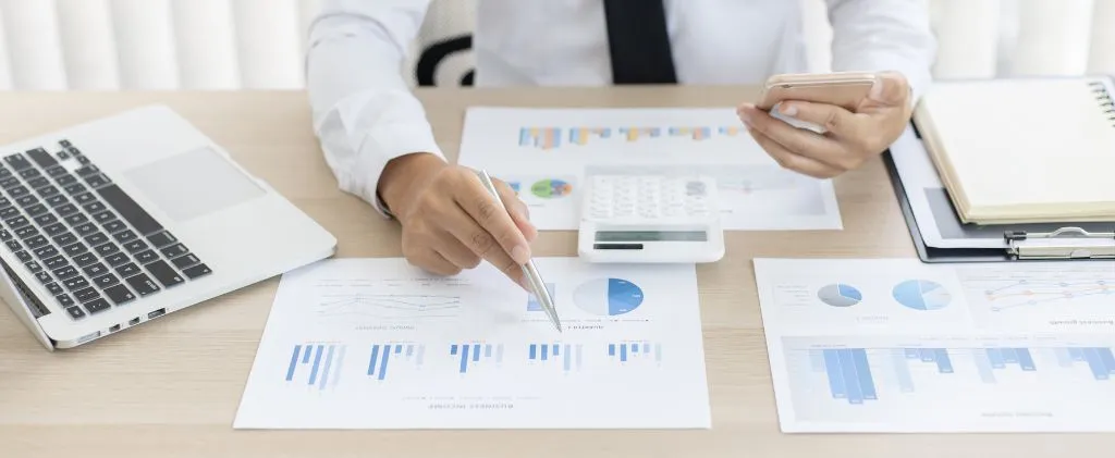 Une personne en tenue de travail examine des graphiques financiers à l'aide d'un stylo, d'une calculatrice, d'un smartphone, d'un ordinateur portable et de documents TRVE Février 2026 posés sur un bureau.